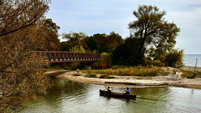 Two canoeists passing a sandy shore and footbridge beside a lake